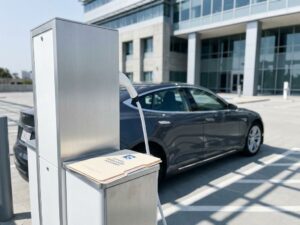Elektrische auto wordt opgeladen bij moderne laadpaal met witte kabel, documenten op station, kantoorgebouw op achtergrond