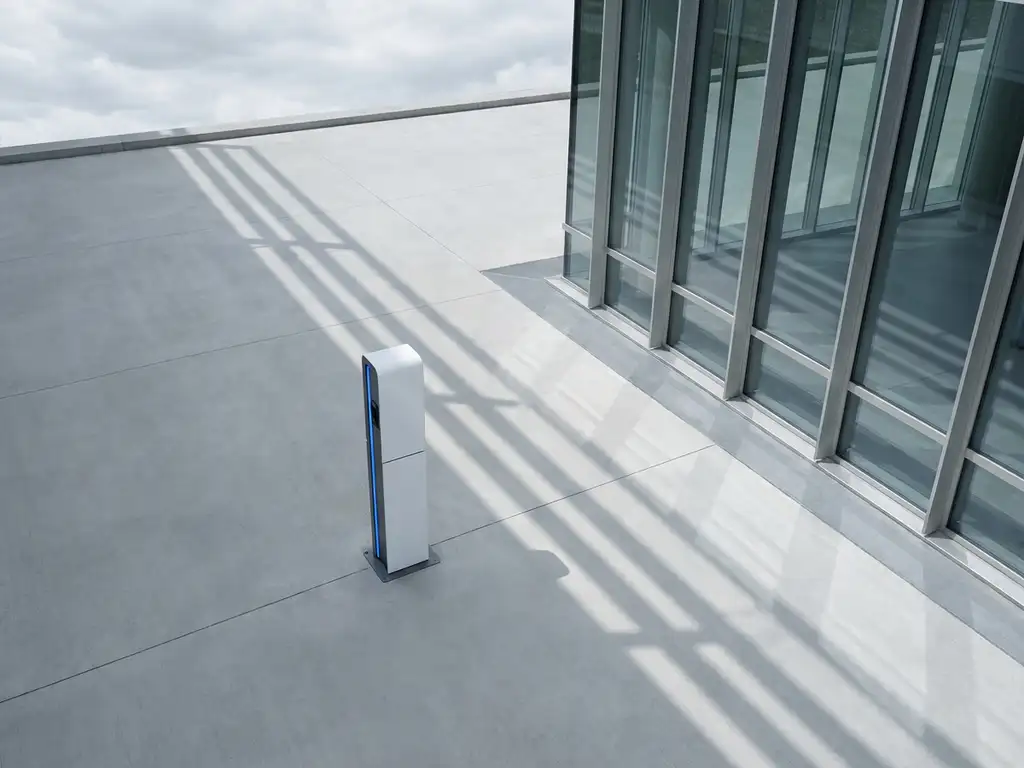 Elektrische laadpaal met wit behuizing en blauwe accenten naast modern glazen kantoorgebouw, geometrische schaduwen op beton